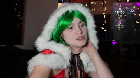 Media: Video of a fair-skinned woman with vibrant green hair in a Santa hat, wearing a red velvet cape. She looks contemplative with one hand on her cheek, set against a dimly lit room with warm, bokeh lights in the background.