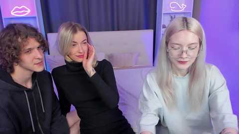 Media: Video of three young adults in a modern bedroom with purple lighting. A blonde woman in a black turtleneck sits, a curly-haired man in a black hoodie stands, and a blonde woman with glasses sits.