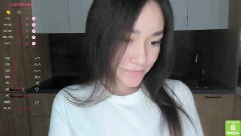 Media: Video of a young Asian woman with long black hair, wearing a white shirt, smiling in a modern kitchen with light wooden cabinets and a dark countertop.