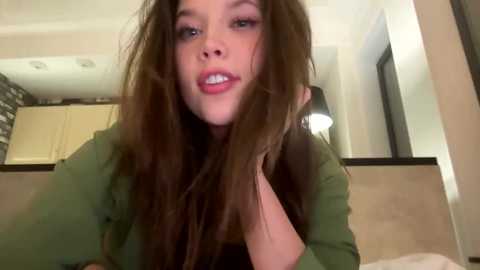 Media: Video of a young woman with long brown hair, pale skin, and full lips, wearing a green top, standing in a modern kitchen with light-colored cabinets.