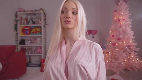 Media: Video of a blonde woman with straight hair wearing a pink satin robe, standing in a festive room with a decorated Christmas tree and a red sofa.