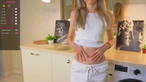 Media: Video of a slender, fair-skinned woman in white crop top and shorts, standing in a modern, light-colored bathroom with white cabinets, a sink, and a washing machine.