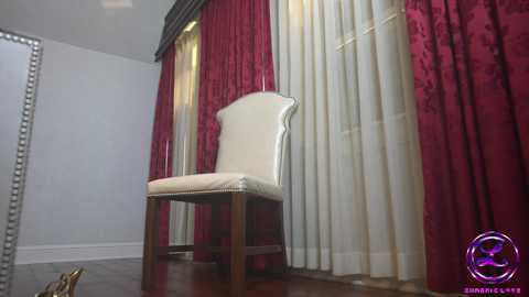 Media: Video of a chic, cream-colored upholstered chair with a wooden frame, set against rich, maroon and white patterned curtains in a spacious, elegant room.