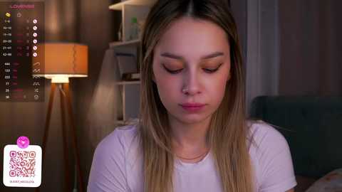 Media: Video of a young, fair-skinned woman with long, blonde hair, wearing a white shirt, looking down. Background includes a lamp and a shelf with books, in a dimly-lit room.