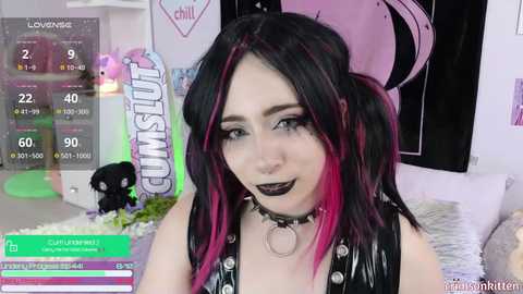 Media: Video of a young woman with black and pink hair, wearing a black choker and a black top, in a pastel-themed room with gaming elements.