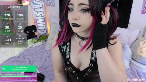 Media: Video of a pale-skinned woman with dark hair, wearing black lipstick and a black leather choker, sitting on a bed with a furry blanket. Background shows a live stream display and plush toys.