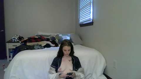 Media: A video of a young woman with long brown hair, wearing a striped shirt, sitting on a bed in a sparsely decorated bedroom, holding a smartphone, with clothes strewn on the bed and a window with closed blinds in the background.