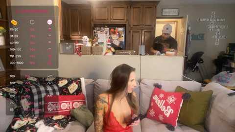 Media: Video of a woman in a red lace lingerie top, tattoos on her arm, sitting on a couch with festive pillows, while a man cooks in the kitchen behind her.