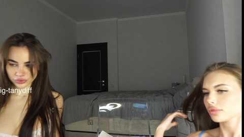 Media: Video of two young women with long, dark hair, one with a phone to her ear, the other with makeup brushes. They stand in a dimly lit, minimalist bedroom with a white bed and a closed door.