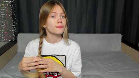 Media: Video of a young Caucasian woman with long blonde hair, wearing a white T-shirt, sitting on a grey bed with black curtains in the background.