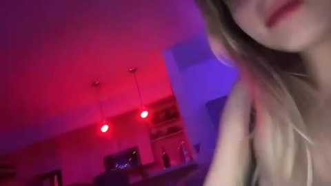 Media: A video featuring a close-up of a blonde woman's face, partially obscured, in a dimly lit room with purple and red lighting. The background shows a kitchen with hanging lights and blurred shelves.