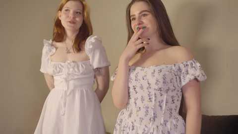 Media: Video of two young women with pale skin, one with long red hair in a white floral dress, the other with long brown hair in a white off-shoulder dress. They are indoors, smiling and holding phones, against a beige wall.