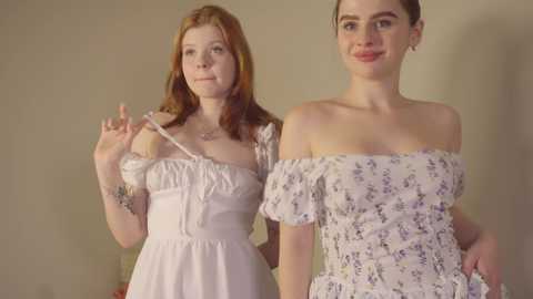 Media: Video of two young women with fair skin, red hair, and freckles, wearing off-shoulder floral dresses. One adjusts her strap, smiling; the other stands beside her, smiling.