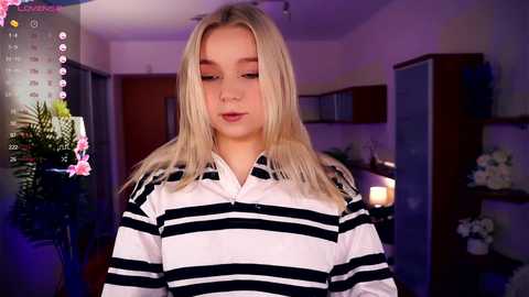 Media: Video of a young blonde woman with fair skin, wearing a black and white striped polo shirt, in a modern kitchen with purple lighting, potted plants, and a calendar overlay.