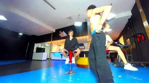 Media: Video of a mixed martial arts gym featuring two men grappling; one is shirtless, the other in a black hoodie, on a blue mat floor, with training equipment and a TV screen in the background.