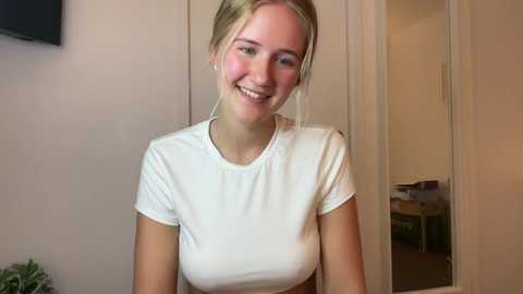 Media: Video of a smiling, fair-skinned young woman with blonde hair tied back, wearing a white t-shirt. Background shows a beige wall, TV, and open doorway.