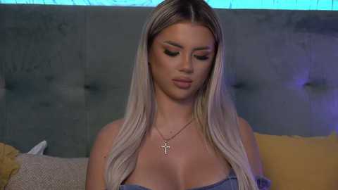 Media: Video of a blonde woman with long, straight hair, wearing a strapless dress, and a cross necklace. She has fair skin, full lips, and subtle makeup. Background features a gray upholstered headboard and a yellow pillow.