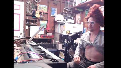 Media: Video of a woman with red hair in glasses, playing a synthesizer in a cluttered, colorful room with various musical instruments and art on the walls.