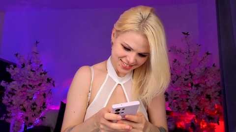 Media: Video of a smiling, fair-skinned, blonde woman with straight hair wearing a white halter top, holding a smartphone. Background features purple lighting, cherry blossom branches, and blurred furniture.