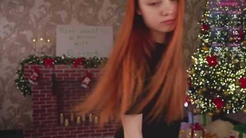 Media: Video of a young woman with long, bright orange hair, wearing a black top, standing in a festive room with a decorated Christmas tree, brick fireplace, and a chalkboard sign.