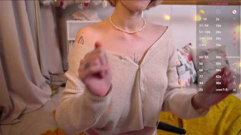 Media: A video shows a woman with short hair, fair skin, and a tattoo on her shoulder, wearing a beige off-shoulder sweater and a pearl necklace, gesturing with her hands in a cozy, dimly lit room with a yellow blanket and a digital display in the background.