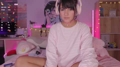 Media: Video of a young woman with fair skin, dark hair, and large headphones, wearing a pink striped sweater, sitting on a bed with stuffed toys and pink bedding. The room is decorated with anime posters and fairy lights, creating a cozy, playful atmosphere.