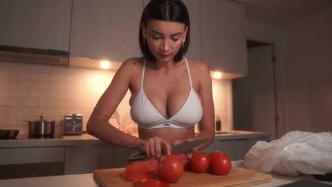 Media: Video of a woman with straight black hair and fair skin, wearing a white bra, chopping tomatoes in a modern kitchen with white cabinets and a tiled backsplash.