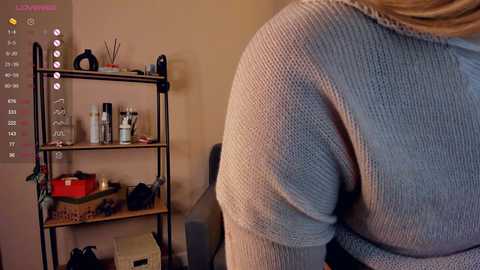 Media: Video of a woman's backside in a tight, light blue knitted sweater. She stands in a room with beige walls, a black metal shelving unit filled with beauty products, and a chair.