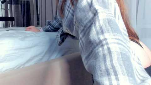 Media: Video of a person with long, straight, light brown hair wearing a grey plaid shirt and black underwear, leaning over a white bed in a modern, brightly lit bedroom.