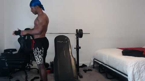 Media: Video of a muscular, shirtless Black man with a blue cap and black patterned shorts lifting weights in a minimalist, white-walled bedroom with a bed and workout equipment.