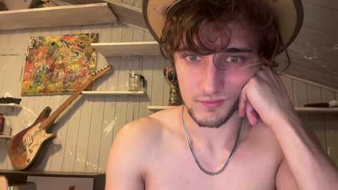 Media: Video of a shirtless young man with curly brown hair and glasses, resting his chin on his hand, wearing a cowboy hat and silver necklace. Background includes a colorful painting, guitar, and shelves in a rustic room.