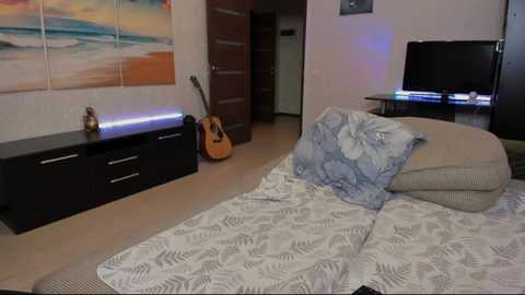 Media: Video of a modern bedroom with a black dresser, guitar, TV, floral-patterned bedspread, and abstract ocean waves wall art.