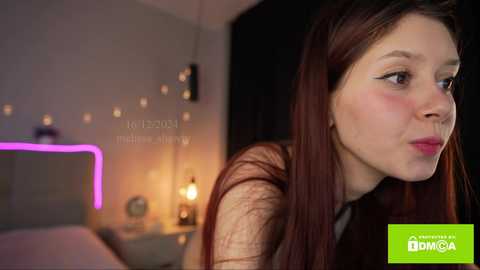 Media: Video of a young woman with fair skin and long brown hair, wearing a sheer black top, looking contemplative in a dimly lit bedroom with a neon pink light, a bed, and a nightstand with a lamp.