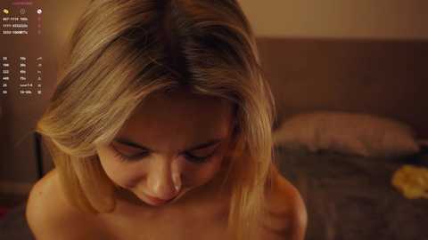 Media: Video of a topless, fair-skinned, blonde woman with medium-length hair, looking down. She's in a dimly lit bedroom with a bed in the background.