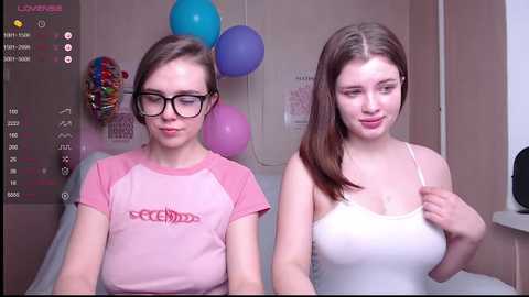 Media: Video of two young women, one with glasses and a pink shirt, the other with a white tank top, in a room with colorful balloons and a poster.