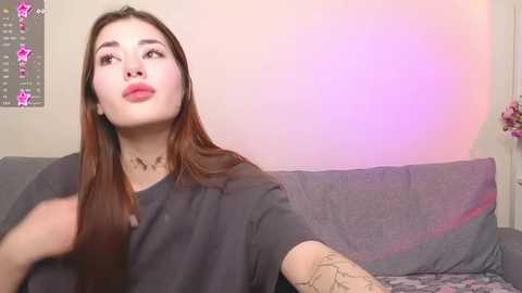 Media: Video of a young woman with long, straight brown hair, fair skin, and full red lips, wearing a dark grey T-shirt, sitting on a grey couch. Pink star filters overlay the image.