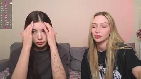 Media: Video of two young women with long hair, one with hands on head, the other with hands on hips, in a cozy living room with a gray couch and pink wall.