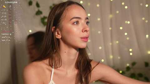 Media: Video of a young woman with light skin, long brown hair, and a slender physique, wearing a white spaghetti-strap top, looking thoughtful. Background features soft, glowing fairy lights and green foliage.