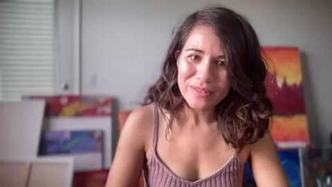 Media: Video of a smiling young woman with wavy brown hair, wearing a sleeveless, ribbed top, in an art studio with colorful abstract paintings on the wall.