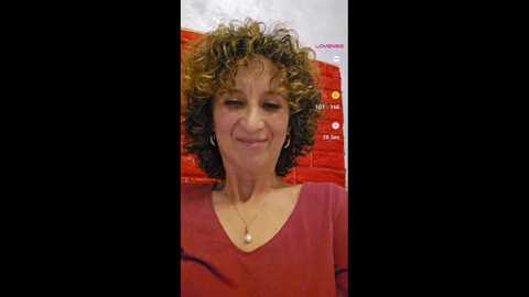 Media: Video of a middle-aged woman with curly brown hair and a pink top, smiling. Background features a red and white advertisement.