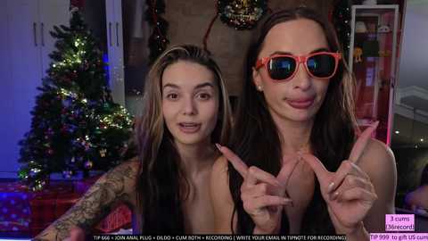 Media: Video of two young women, one topless with tattoos, wearing red sunglasses, and the other smiling, in a festive, decorated living room.