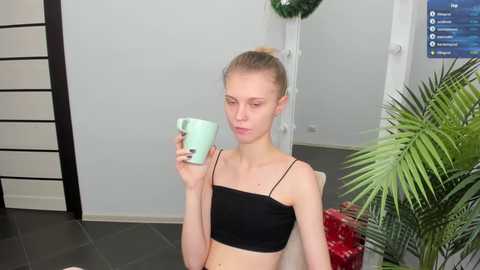 Media: Video of a slender, fair-skinned woman with blonde hair in a bun, wearing a black tank top, holding a mint green mug, sitting in a modern, minimalist room with a potted plant and holiday decorations.