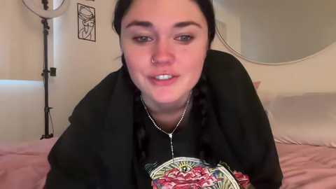 Media: Video of a young woman with fair skin, dark hair, and a nose ring, wearing a black graphic tee, lying on a pink bed, with a round mirror and lighting equipment in the background.