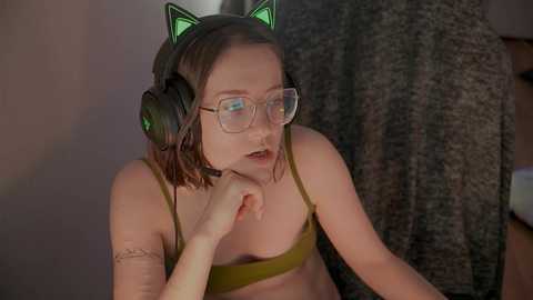 Media: Video of a young woman with glasses, green cat ears, and headphones, wearing a green bra, sitting in a dimly lit room, with a textured curtain and wooden chair in the background.