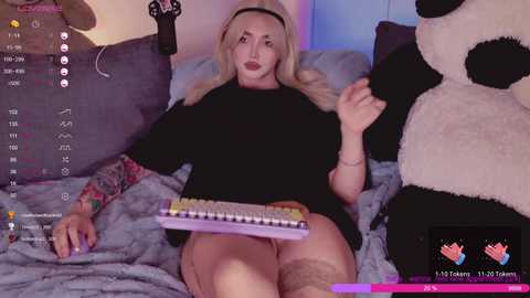 Media: Video of a blonde woman with a black headband, wearing a black top, playing a keyboard on a bed with plush toys.