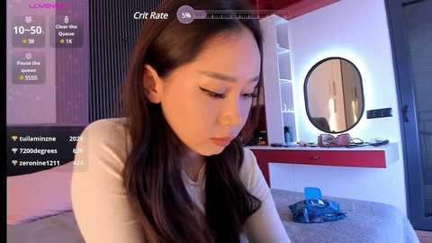 Media: A video of an Asian woman with long brown hair, wearing a white top, sitting in a modern bedroom with a round mirror, digital clock, and a blue toy.