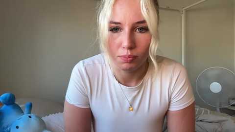 Media: Video of a blonde woman with a pouty face, wearing a white T-shirt, sitting on a bed in a dimly lit room. Background includes a stuffed toy and a fan.