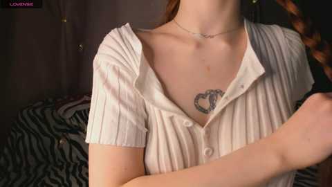 Video of a fair-skinned woman with a heart tattoo on her collarbone, wearing a partially unbuttoned white ribbed shirt, revealing her cleavage. Background includes zebra-print fabric and dim lighting.