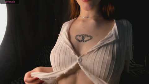 A video of a fair-skinned woman with red hair, wearing a semi-open white ribbed sweater, revealing a tattoo of a heart with a key on her chest.