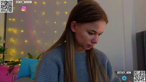 Media: Video of a young woman with long, straight brown hair, wearing a gray sweater, seated indoors, looking down, with a blue and pink background, featuring fairy lights.